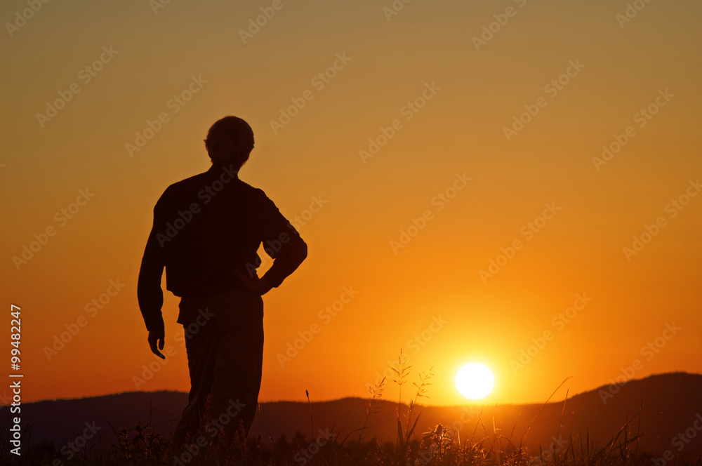 Man watching the sunset on a grassy horizon. Silhouette. Wooded mountains in the background.