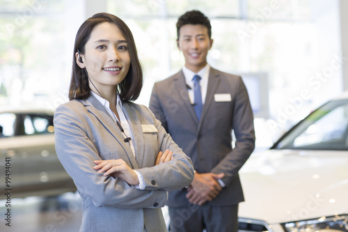 Confident salespeople standing with new cars in showroom