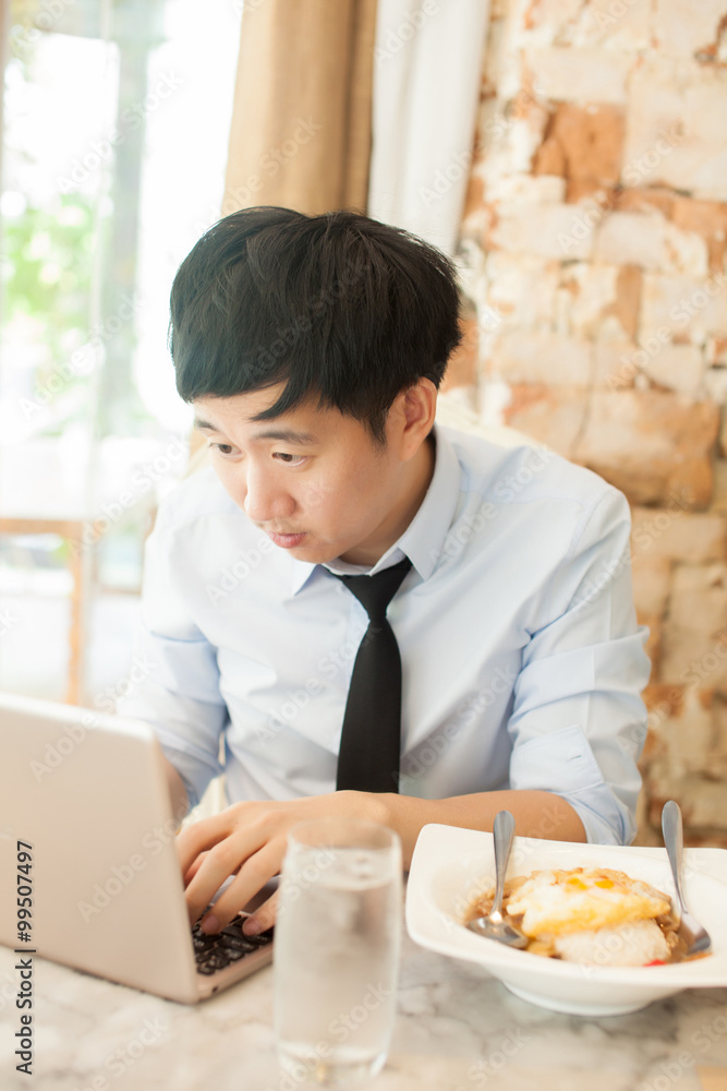 Young Asian man working while eating with his laptop in restaurant (selective focus)