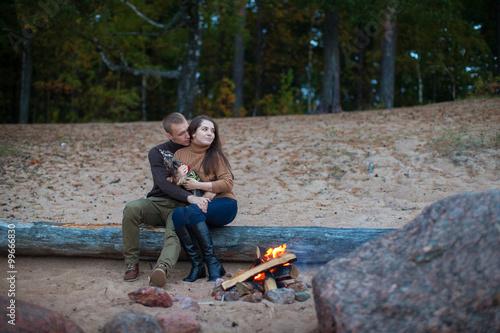 Loving couple with dog sitting on a log by the fire on the beach