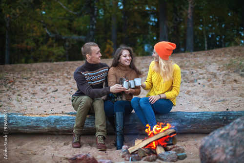 small company communicates sitting on a log by the fire on the beach against the background of the forest