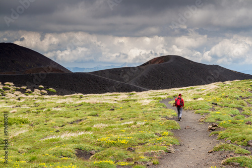 Lonely unrecognizable Hiker dressed in in red walks along a path on Mount Etna in Sicily, heading to a black volcano crater.
