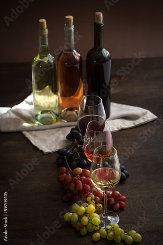 Red, rose and white wine bottles with three wineglasses and grapes on brown wood textured table covered with canvas towel