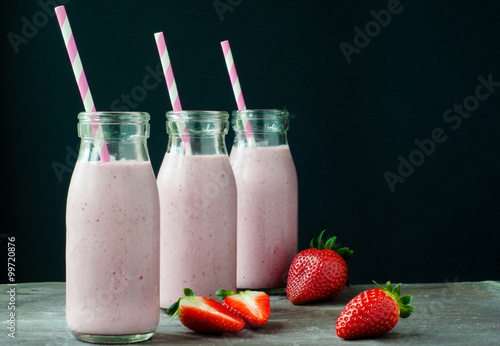 Smoothie milkshake made from fresh strawberry blended with kefir yogurt. Served in bottle style glasses and with fresh strawberries. Served on a stone slate table with colorful stripy straws.