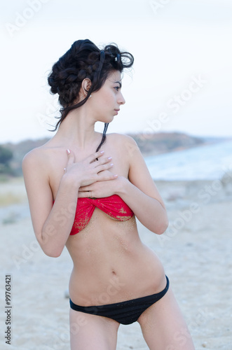 Young slim pretty lady wear red and black bikini standing on the beach looking away, hands on her chest
