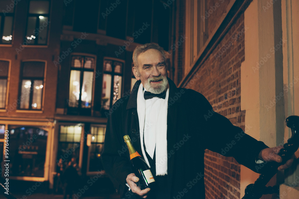 Smiling man in tuxedo with bottle of champagne at door at night. Smiling man in tuxedo with bottle of champagne at door at night.