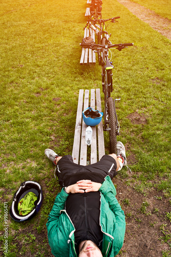 Cyclist resting on a bench in the park. Tired young man with his mountain bike in the forest resting and making pause.