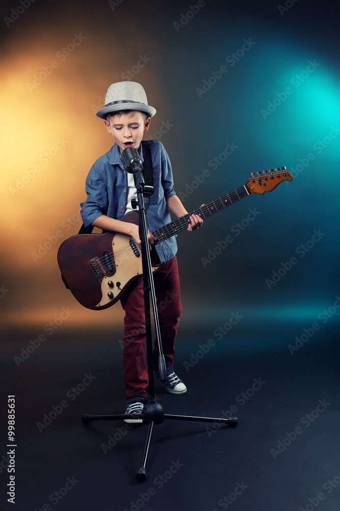 Naklejka premium Little boy playing guitar and singing with microphone on a dark lighted background