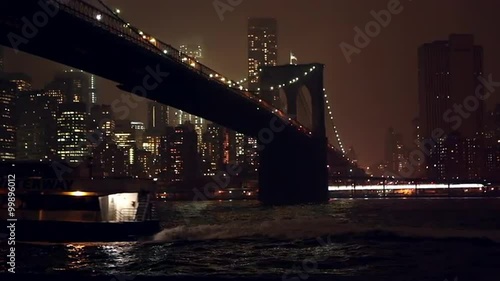 Shot at night overlooking the East River and the Brooklyn Bridge as a boat crusies by.