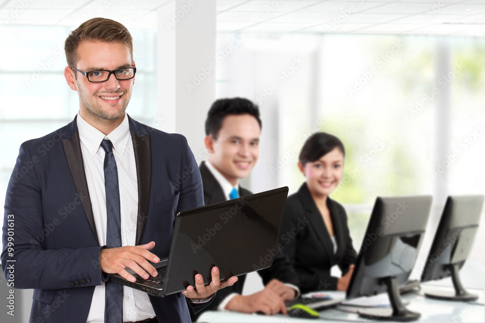 Young caucasian businessman, with his team behind holding laptop Young caucasian businessman, with his team behind holding laptop
