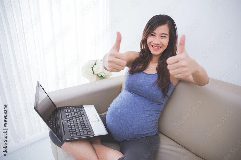 Beautiful pregnant asian woman sitting on a sofa with laptop on