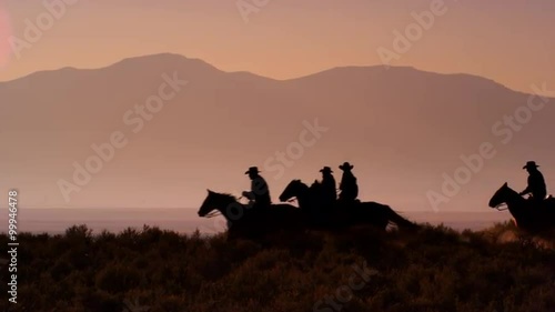 Cowboys galloping riding horses silhouetted against orange mountains in slow motion.