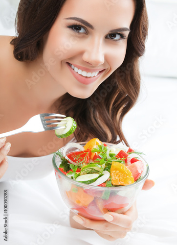 smiling young woman eating salad, on bed