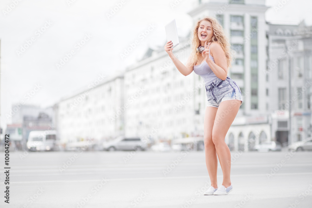 Obraz premium Music and selfie / Happy young woman with vintage music headphones around her neck, taking selfie with tablet pc and smiling happily against urban city background.