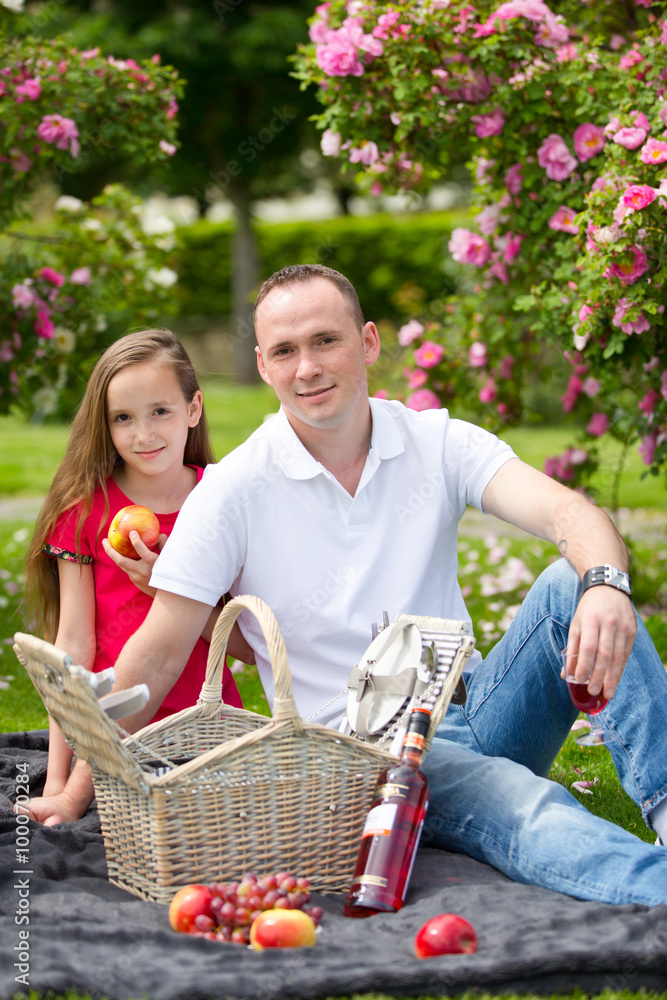 Fototapeta premium Beautiful young father sitting on a plaid in a green park with her small pretty daughter with a wicker basket for happy picnic