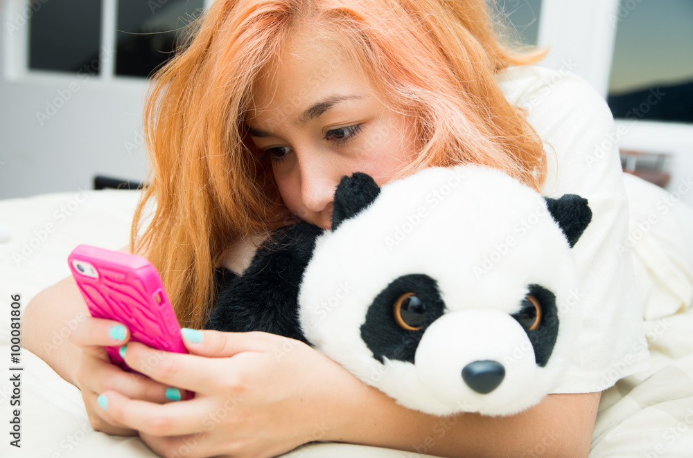 Pretty young woman horizontally lying cozy in bed using pink mobile phone and stuffed panda animal Pretty young woman horizontally lying cozy in bed using pink mobile phone and stuffed panda animal