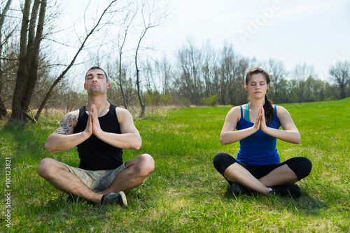 Young man together with young woman is doing yoga exercises on grass .