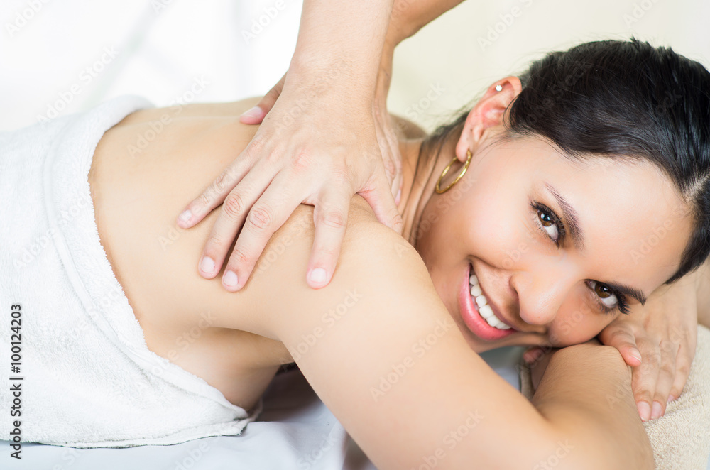 Hispanic brunette model getting massage spa treatment, woman lying upon stomach smiling to camera while hands working on shoulders Hispanic brunette model getting massage spa treatment, woman lying upon stomach smiling to camera while hands working on shoulders
