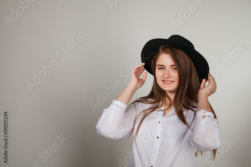 Awesome Caucasian attractive shy sexy female model with brunette hair posing on table in studio, wearing formal suit and hat, isolated on white background