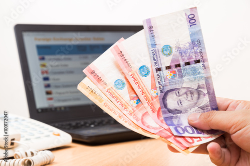 Hand holding Philippines Piso in office with exchange rate on computer screen