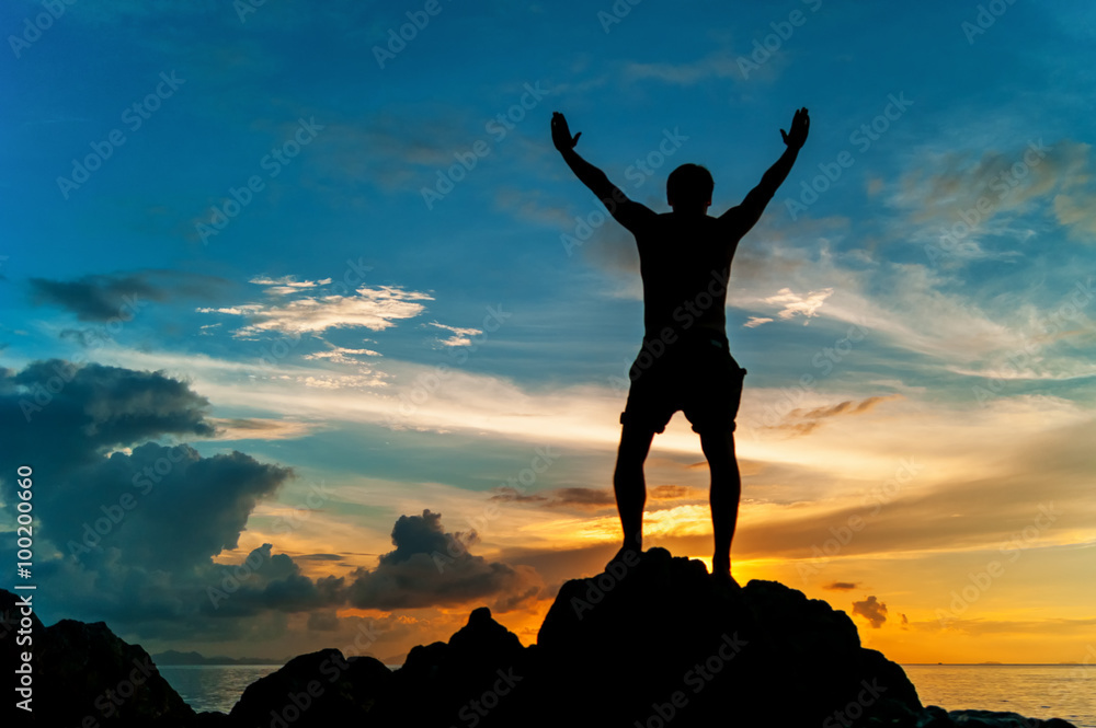 Man standing on the rock of a tropical island with his hands up and watching the sunset