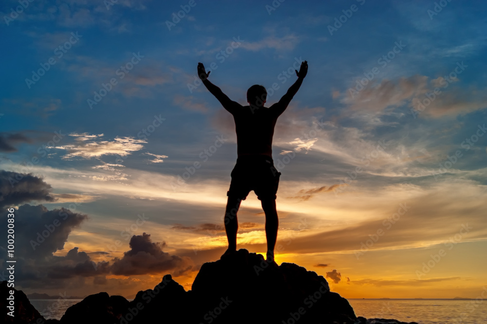 Man standing on the rock of a tropical island with his hands up and watching the sunset Man standing on the rock of a tropical island with his hands up and watching the sunset