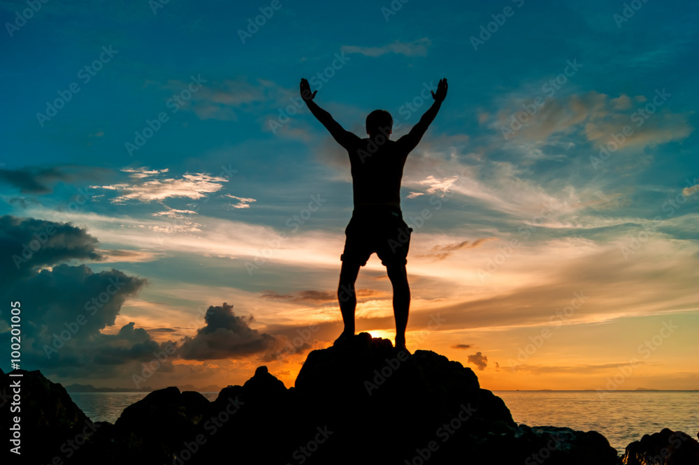 Man standing on the rock of a tropical island with his hands up and watching the sunset