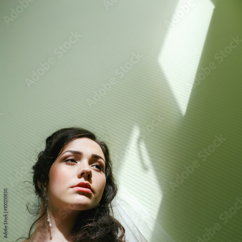Beautiful young woman headshot, professional makeup and hair on green-cyan corrugated wall with patches of light from the windows, tight framing, copy space for text. Thoughtful expression. photo
