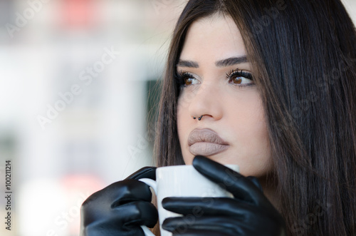 Woman with a unique fleshy lips wearing leather jacket and gloves holding a coffee cup, sitting in cafeteria