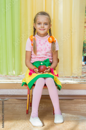 Girl sitting on a chair with an apple in his hand on a matinee in kindergarten