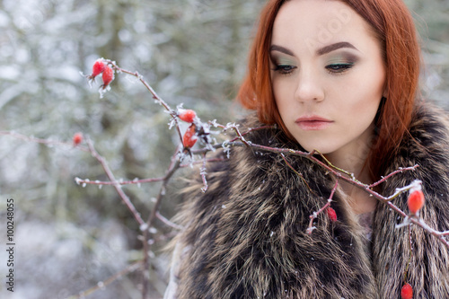 beautiful cute sexy young girl with red hair walking in a snowy forest among the trees missed first trimester bushes with red yagodamiv warm coat with bright makeup photo