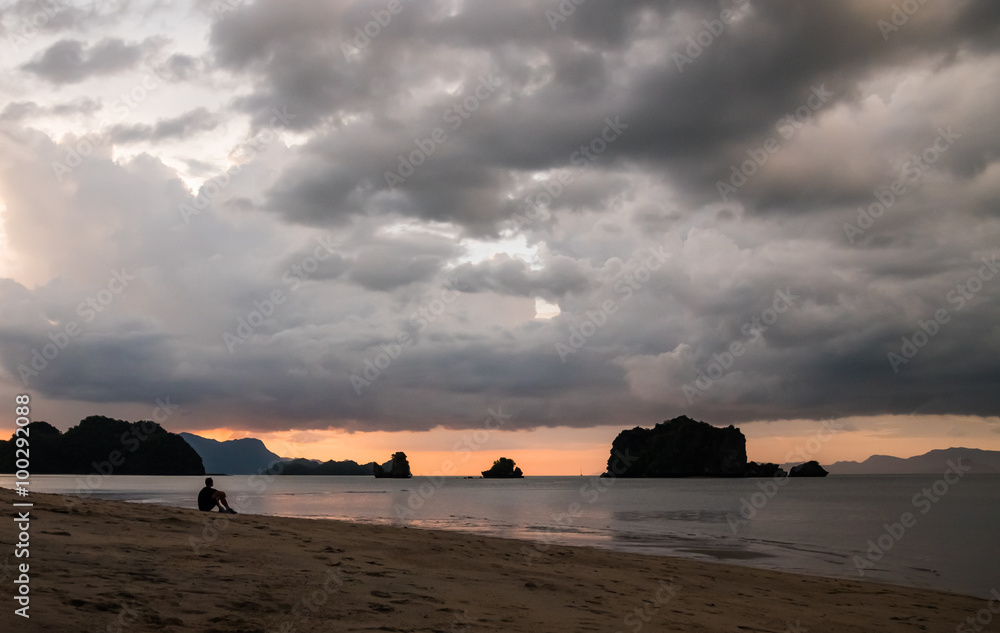 A silhouette of a man is sitting on a tropical beach right after sunset, enjoying the orange sky. Location: Tanjung Rhu Beach, Langkawi, Malaysia.
