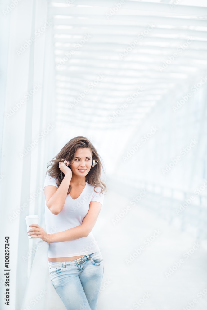 Peaceful morning / Happy young woman wearing music headphones, standing on the bridge with a take away coffee cup and looking aside against urban background Peaceful morning / Happy young woman wearing music headphones, standing on the bridge with a take away coffee cup and looking aside against urban background