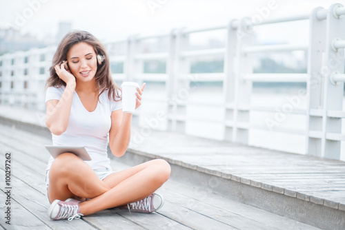 Music and peace / Happy young woman with music headphones and a take away coffee cup, surfing internet on tablet pc, listening to the music and sitting on the bridge.