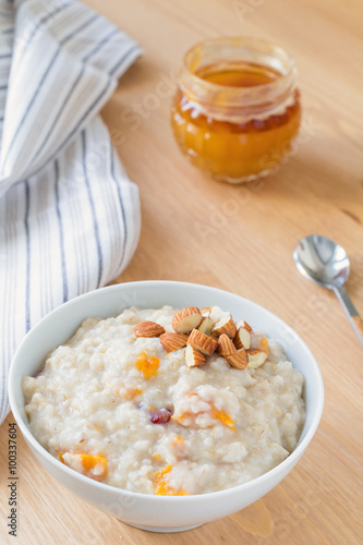 Oatmeal porridge with dried apricots and chopped almonds on bright wooden table. Jar of honey on background. Selective focus. Bright healthy breakfast image
