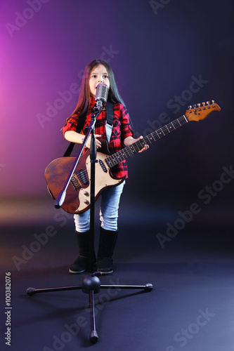 Beautiful little girl with microphone and guitar on dark purple background