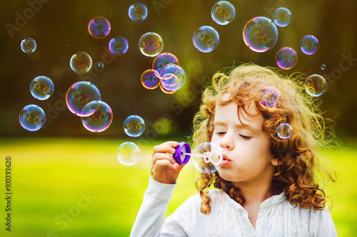 Canvas-taulu Curly little girl blowing soap bubbles in summer park.