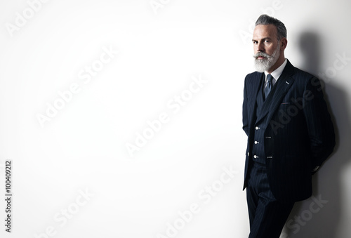 Portrait of bearded businessman wearing trendy suit and stands against the empty white wall. Wide.