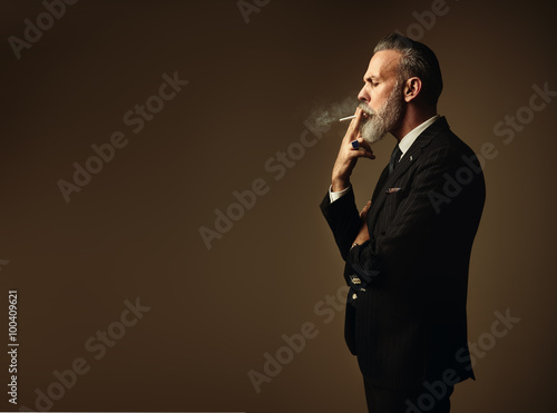 Portrait of smoking gentleman wearing trendy suit and stands against the empty wall. Horizontal photo
