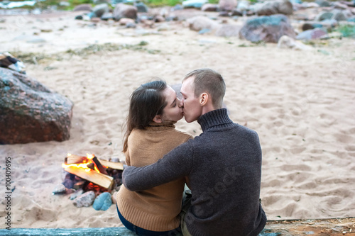 Loving couple sitting on a log by the fire on the rocky beach