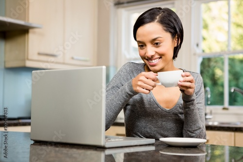 Smiling brunette holding cup and looking at laptop