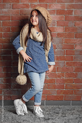 Portrait of little fashion kid girl in knitted cap and scarf on bricks wall background