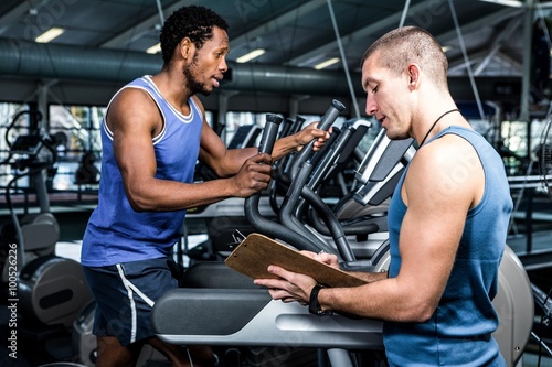 Muscular man using elliptical machine with trainer 