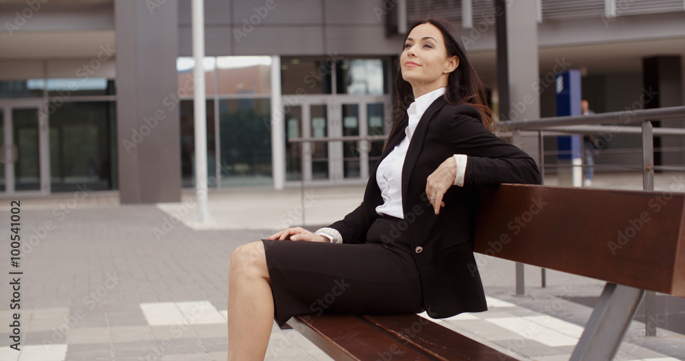 Confident young business woman sitting alone on bench outside office building with calm expression Confident young business woman sitting alone on bench outside office building with calm expression