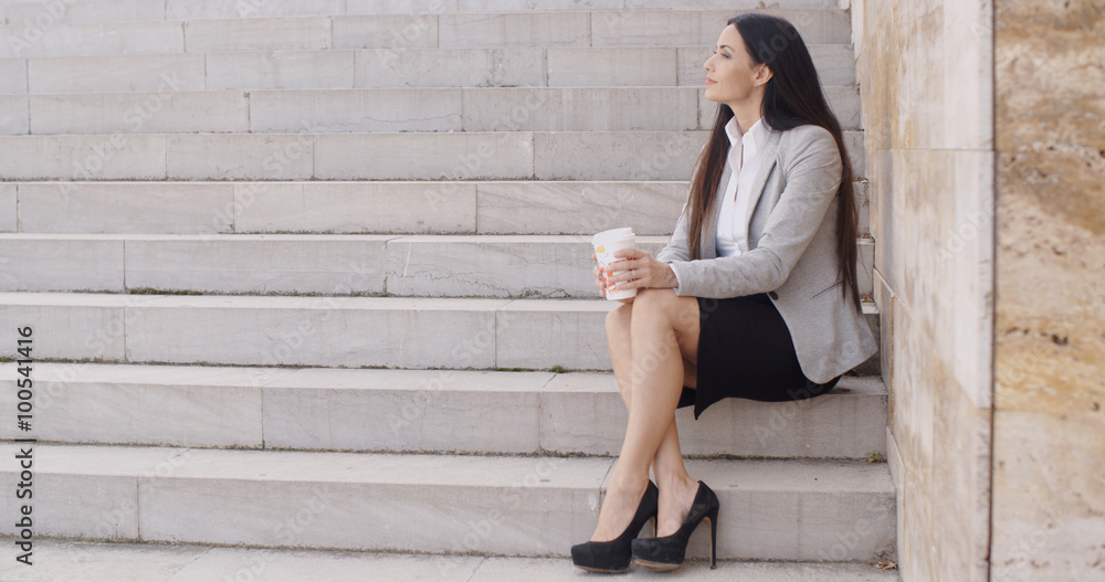 Grinning brunette professional woman sitting on outdoor stairs while drinking coffee