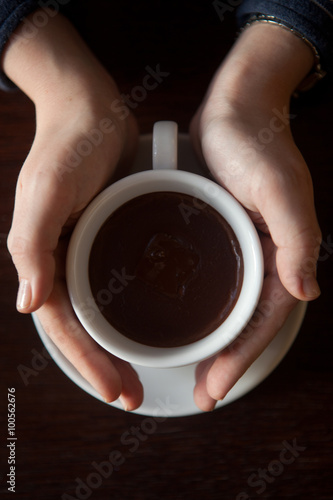 Female hands holding a cup of hot chocolate with foam over wooden table, top view 