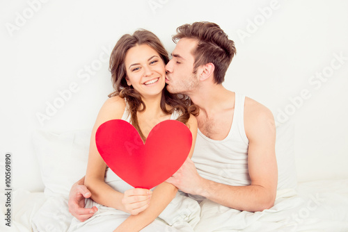 Lovely couple in love holding red paper heart in bed and kissing