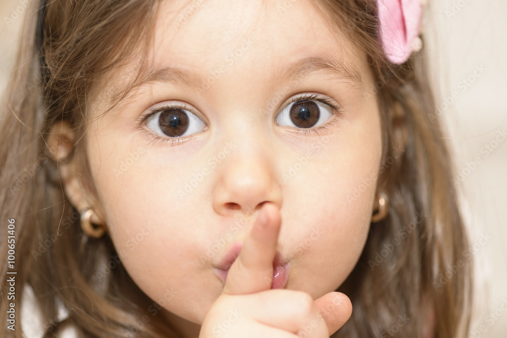 Portrait of expressive beautiful little girl finger up to lips for making a quiet gesture. Girl showing silence gesture looking at the camera Portrait of expressive beautiful little girl finger up to lips for making a quiet gesture. Girl showing silence gesture looking at the camera