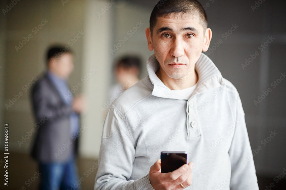 Middle Asian man short haircut with phone in his hand questioning look directly into camera, gets a job. Against background of business men discussing employment. Unemployment, human resources. Middle Asian man short haircut with phone in his hand questioning look directly into camera, gets a job. Against background of business men discussing employment. Unemployment, human resources.