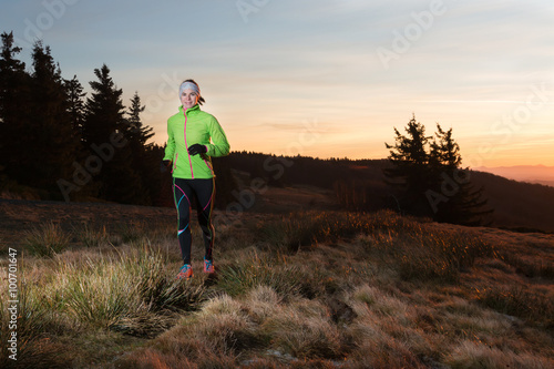 smiling girl in green jacket running on the trail in the hills early morning before sunrise